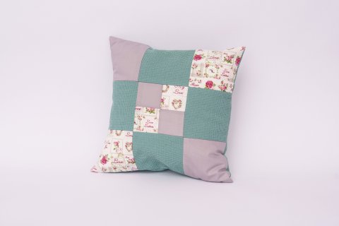 Ein quadratisches Patchwork-Kissen mit einem Muster aus verschiedenen Stoffquadraten liegt vor einem hellen Hintergrund. Die Stoffe sind in Pastellt&ouml;nen gehalten &ndash; unter anderem in Hellgr&uuml;n, Flieder und mit floralen Mustern in Rosa. Das Kissen wirkt handgemacht und dekorativ.