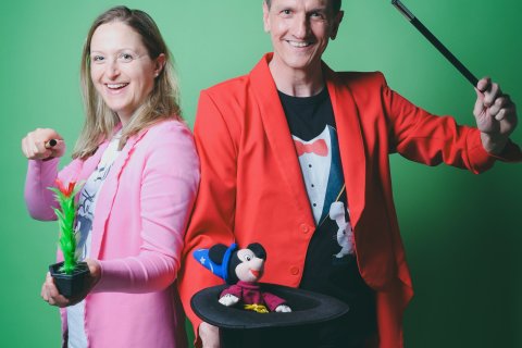 Vor einem gr&uuml;nen Hintergrund stehen eine Frau in pinkem Blazer und ein Mann in Zauberkost&uuml;m mit Zylinder in der Hand. Beide haben einen Zauberstab in der Hand und richten diese in Richtung Betrachter des Bildes.