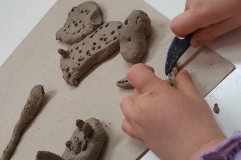 Zu sehen sind zwei Kinderh&auml;nde, die aus Ton auf einer Bastelunterlage Fantasietiere herstellen mit einem kleinen messerartigen Werkzeug wird der Ton bearbeitet
