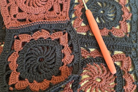 Zu sehen sind geh&auml;kelte bereits zusammengen&auml;hte Granny Squares in den Farben schwarz und rostbraun und eine H&auml;kelnadel mit orangefarbenem Griff.