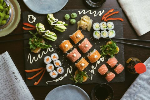 Gro&szlig;e Platte mit verschiedenen Sushi‑Rollen (Lachs und Avocado), dekoriert mit Sesam, Ingwer, Wasabi und So&szlig;en, daneben Edamame, St&auml;bchen und kleine Schalen auf einem Holztisch.
