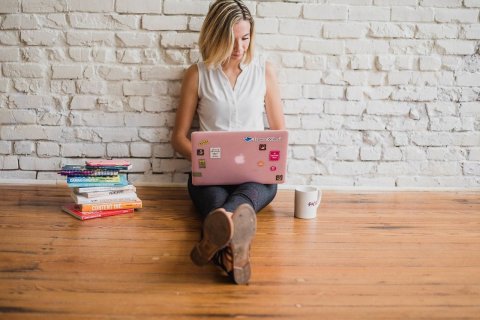 Auf dem Bild sitzt eine Frau auf dem Boden, sie tippt etwas in ihr Laptop.