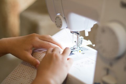 Zu sehen ist eine N&auml;hmaschine die von einem Kind bedient wird wlches gerade einen wei&szlig;en Stoff unter dem N&auml;hfu&szlig; f&uuml;hrt. man sieht nur die Maschine und die H&auml;nde des Kindes.